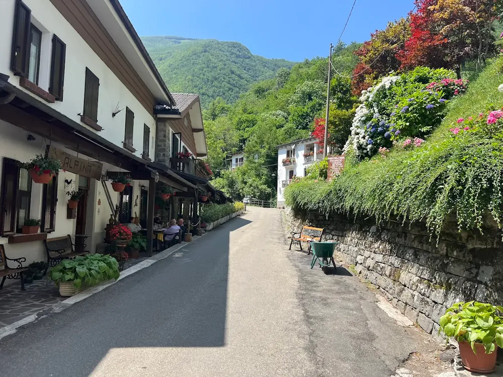Via Roma a Pianaccio: la veranda della Locanda con l'insegna in legno, gerani alle finestre, ortensie e la montagna verde sullo sfondo.