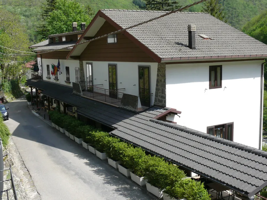 La Locanda vista dall'alto: il tetto scuro, la veranda coperta lungo la strada, le siepi in vaso e il bosco tutto intorno.